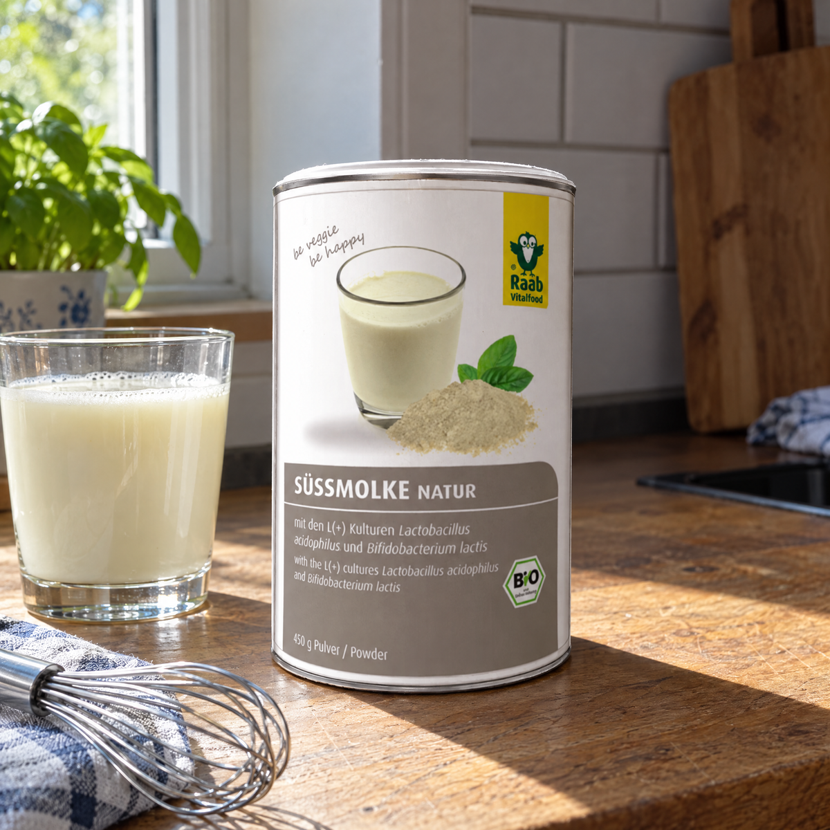 Raab Vitalfood BIO Premium Süssmolke-Pulver Vitaldrink, 450 g – auf der Küchenablage neben einem gefüllten Glas und einem Schwingbesen